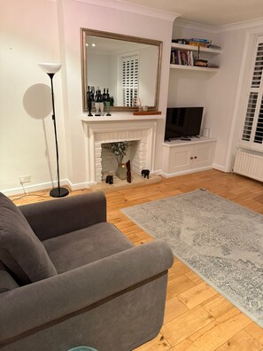 Ein Wohnzimmer mit grauem Sofa, einer Stehlampe, einem Kamin, einem Fernseher und einem Bücherregal voller Gesellschaftsspiele.