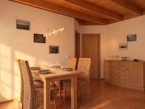 A dining area with a wooden table set for two, surrounded by woven chairs, with a wooden ceiling and a door in the background. - Ferienhaus Fünfseen-Glück (Fünfseen)