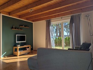 A living room with a wooden ceiling, a television on a wooden stand, a grey sofa, and a sliding glass door leading to a garden. - Ferienhaus Fünfseen-Glück (Fünfseen)