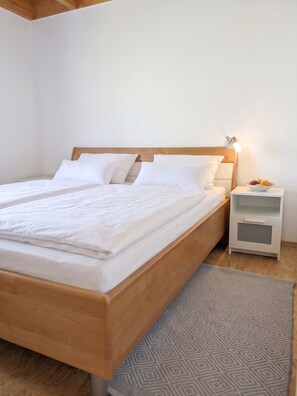 A neatly made bed with white linens, a wooden headboard, a bedside table with a lamp, and a patterned rug. - Ferienhaus Fünfseen-Glück (Fünfseen)