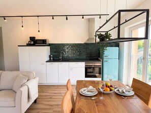 Een moderne keuken met witte kasten, een zwart aanrecht en een groene achterwand. Er is een eethoek met een houten tafel voor twee personen, een bank en uitzicht op het omliggende landschap.