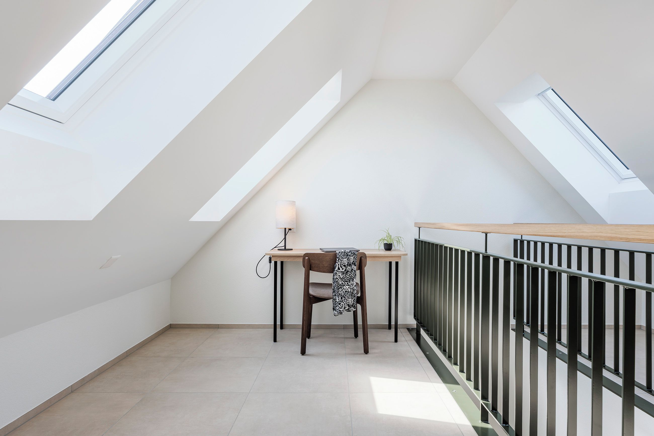 Une petite pièce mansardée, bien éclairée, avec un plafond en pente, un bureau en bois, une chaise, une lampe et une vue à travers un puits de lumière.