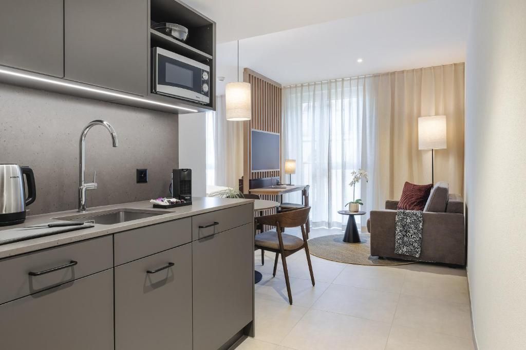 A modern kitchen with a microwave, sink, and coffee maker. There is a dining area with a table and chairs, and a living room with a sofa and a lamp.