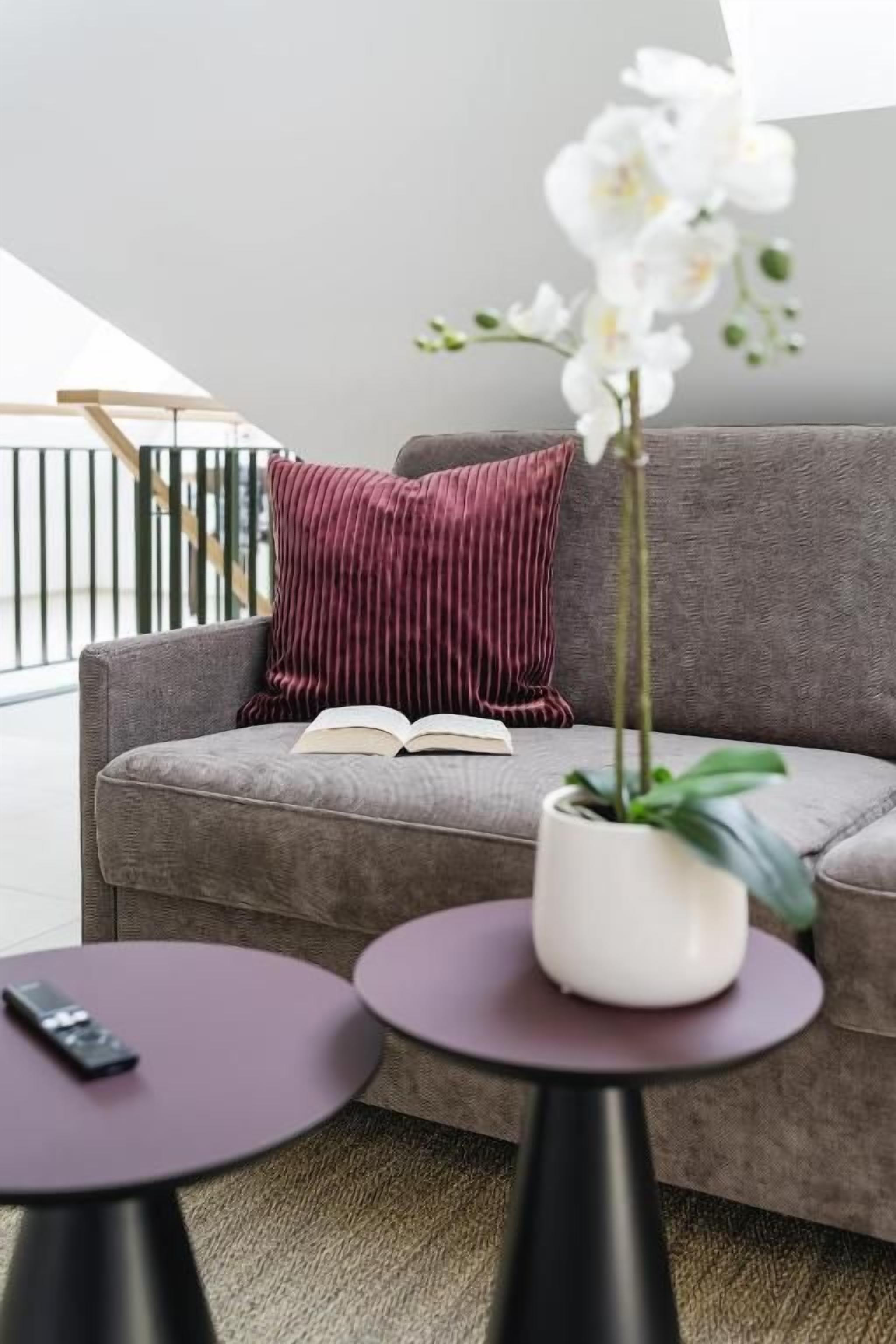 Un salón moderno con un sofá gris, una almohada roja, una orquídea blanca en una maceta y un control remoto sobre una mesa redonda.