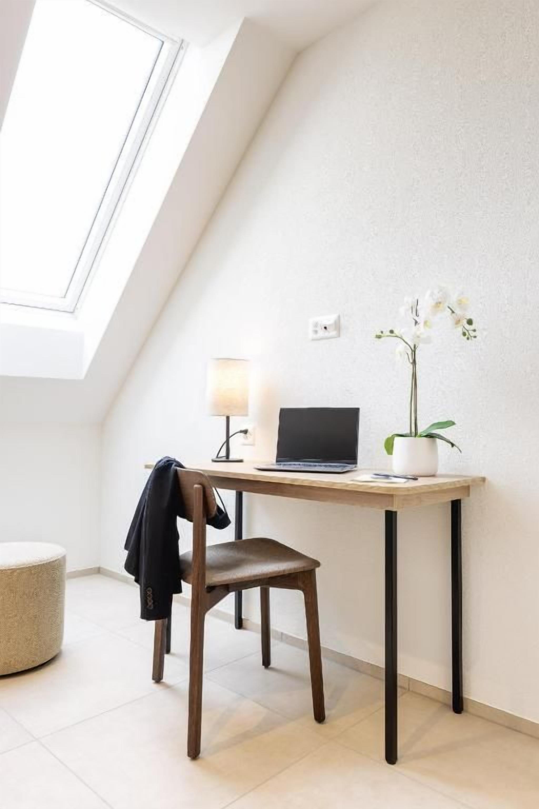 Ein Zimmer mit Schreibtisch, Stuhl, Laptop, Lampe und einem Blumentopf.