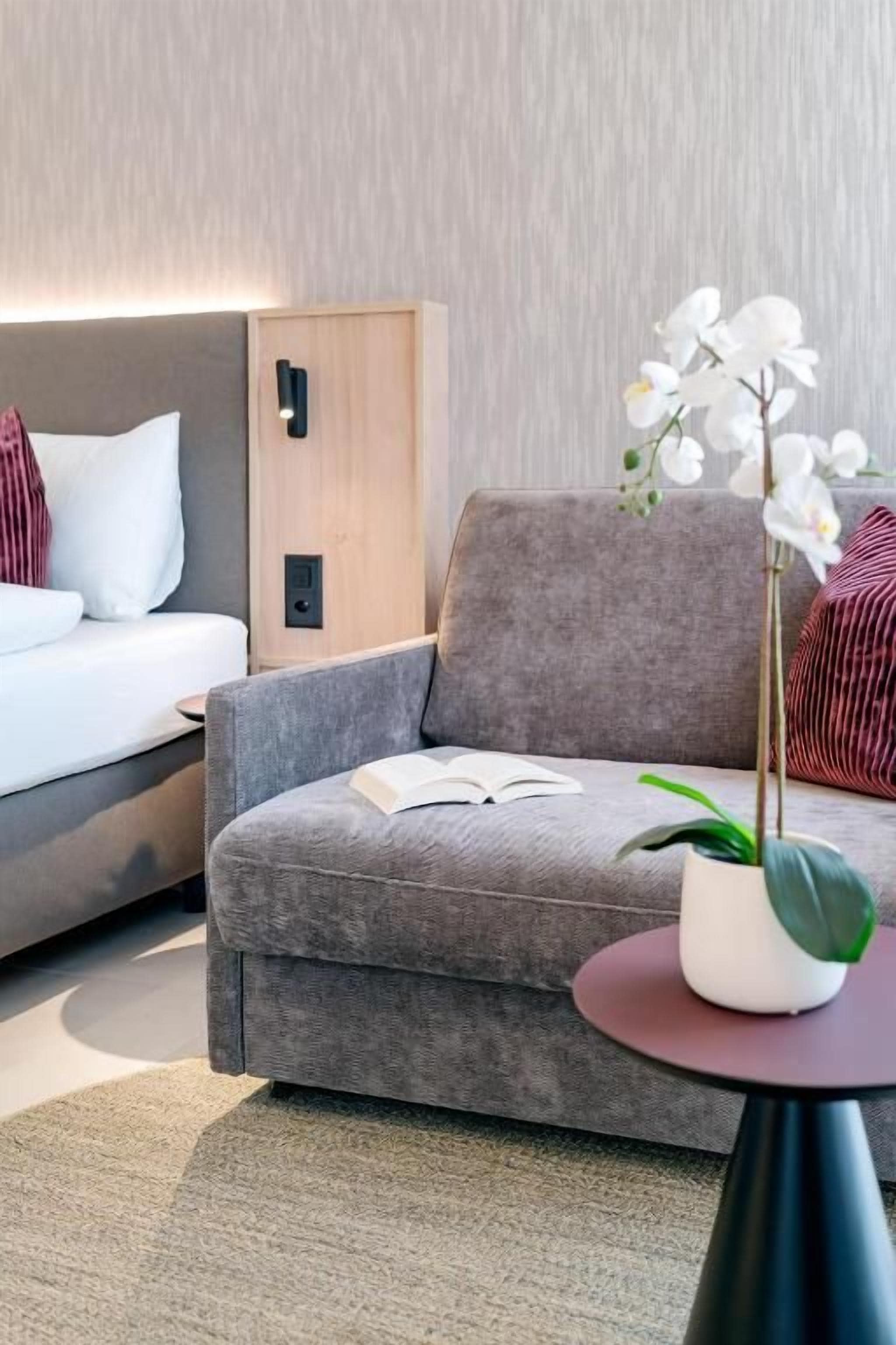 Ein modernes Hotelzimmer mit einem grauen Sofa, einem kleinen runden Tisch mit einer Topfpflanze und einem Bett mit weißer Bettwäsche.