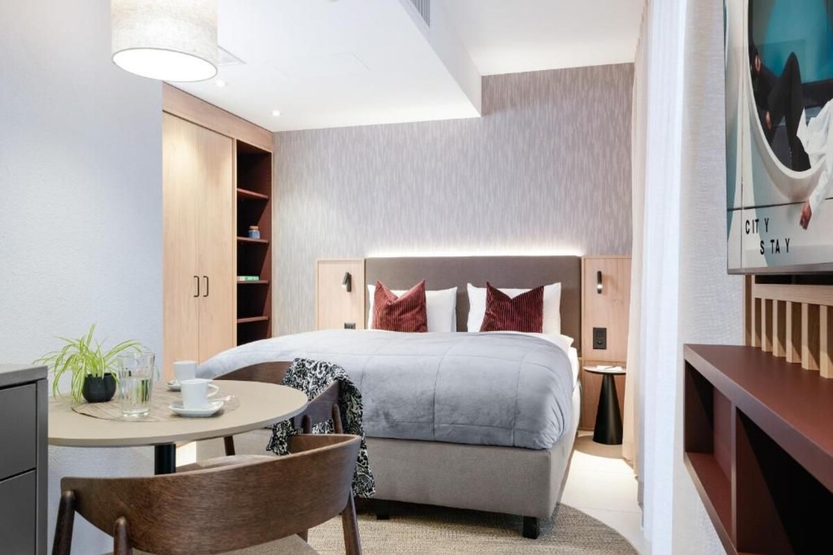 Una habitación de hotel con una cama, mesitas de noche, un escritorio con una silla, una pequeña mesa con una planta y un televisor montado en la pared.