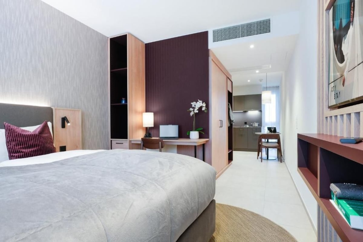 Habitación de hotel moderna con una cama grande, un escritorio, una silla, una mesita y una kitchenette al fondo.