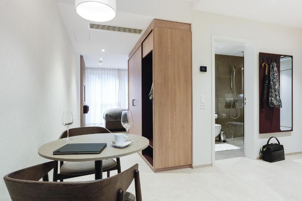 Quarto de hotel moderno com mesa de jantar redonda, duas cadeiras, guarda-roupa, banheiro com box de vidro e espelho com toalha.
