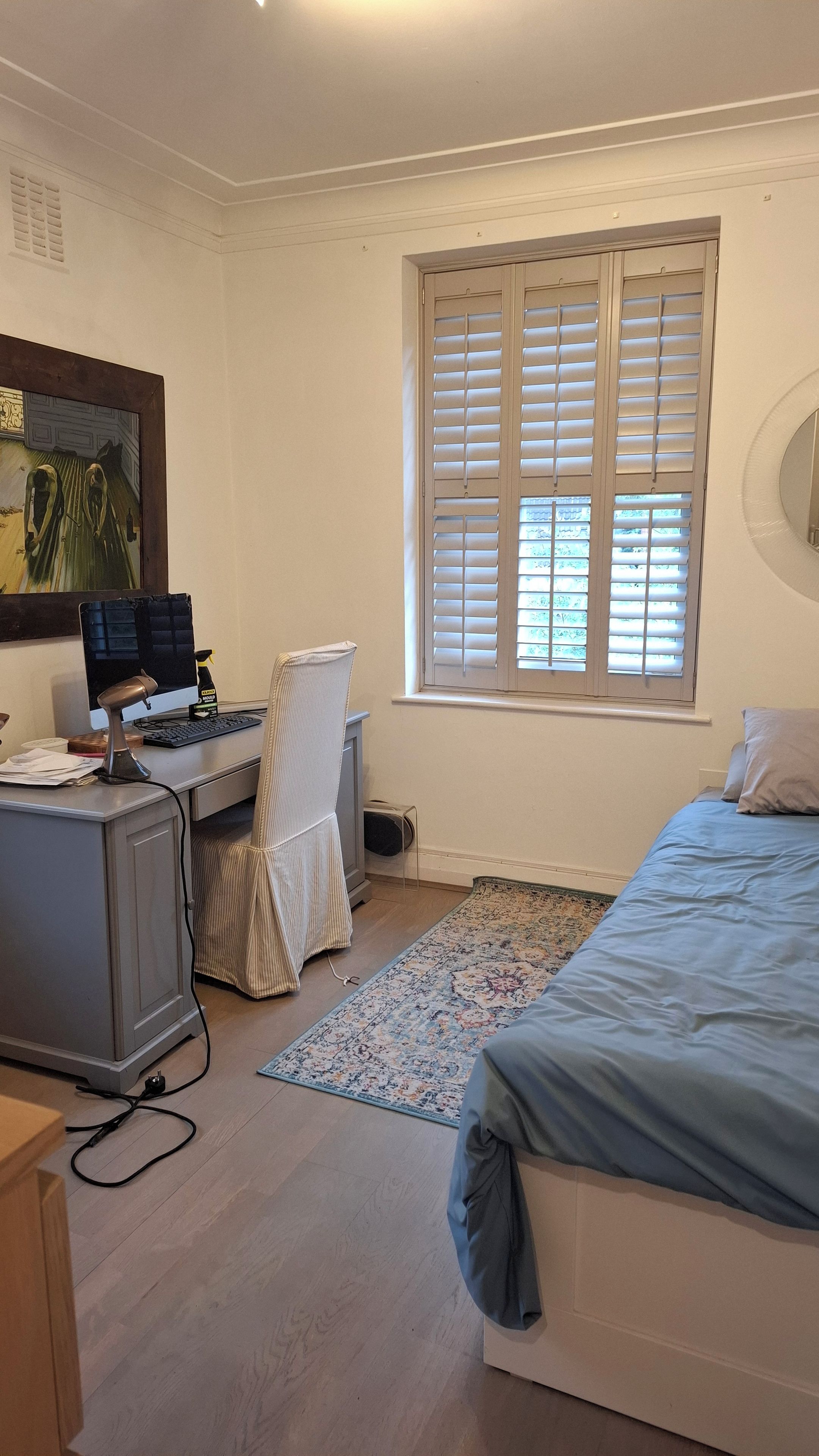 A bedroom with a bed, a desk with a computer, a chair, a window with blinds, and a rug.