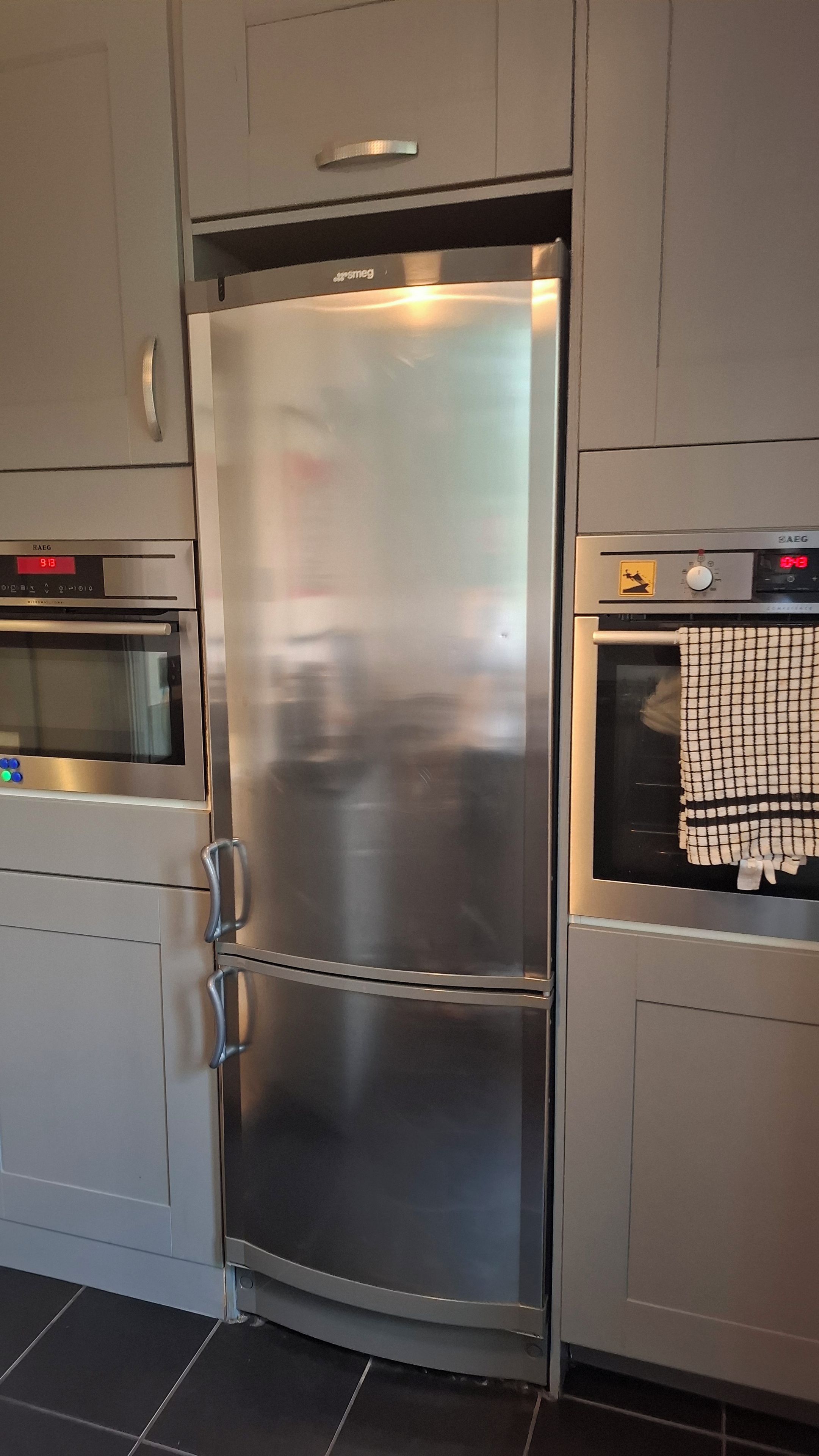 Frigorifero in acciaio inossidabile con doppie ante e display digitale, posizionato tra un forno a microonde e una lavatrice in una cucina.