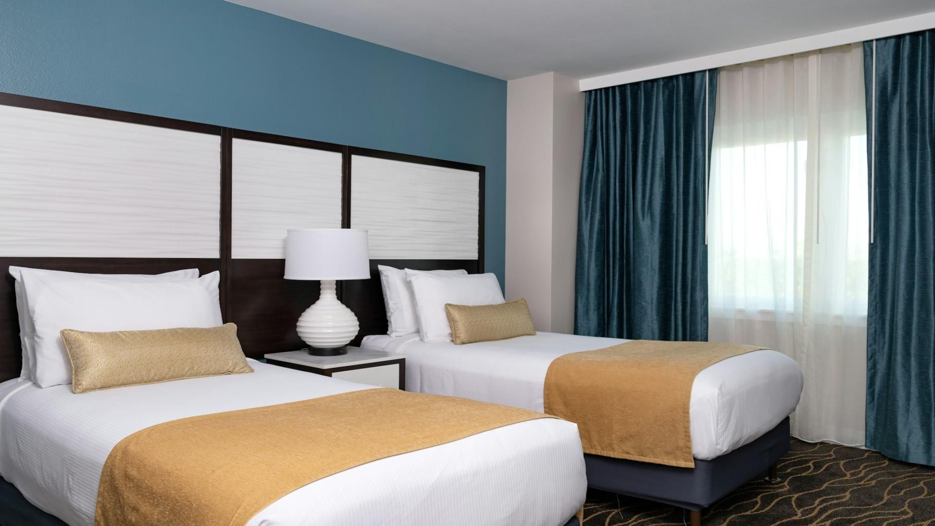 A hotel room with two beds, a nightstand with a lamp, a window with curtains, and a patterned carpet.
