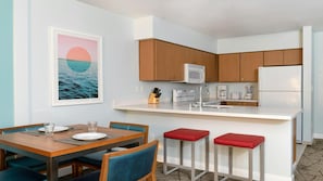 Een moderne keuken met houten kastjes, een wit aanrecht, een roestvrijstalen spoelbak en een koelkast. Er is een eethoek met een houten tafel en blauwe stoelen.