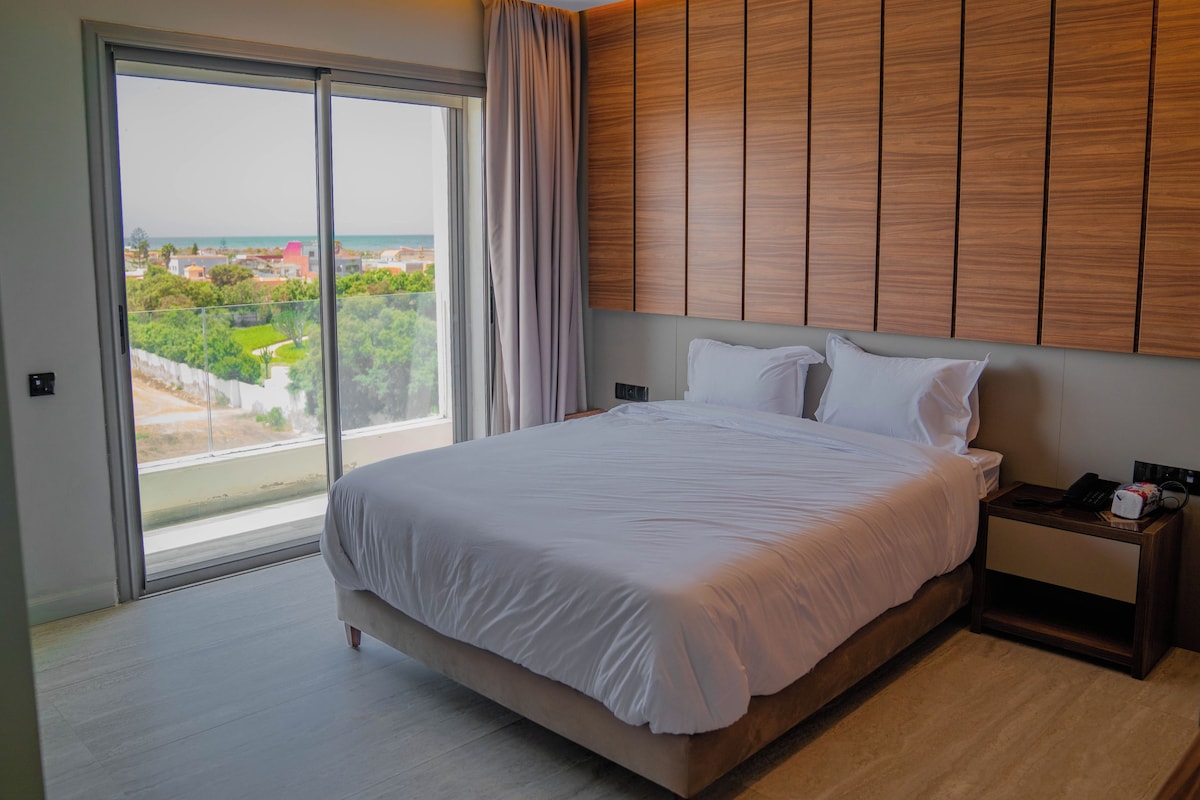 Habitación de hotel con una cama grande, una mesita de noche, un cabecero de madera y vistas a un paisaje urbano a través de una puerta corrediza de cristal.