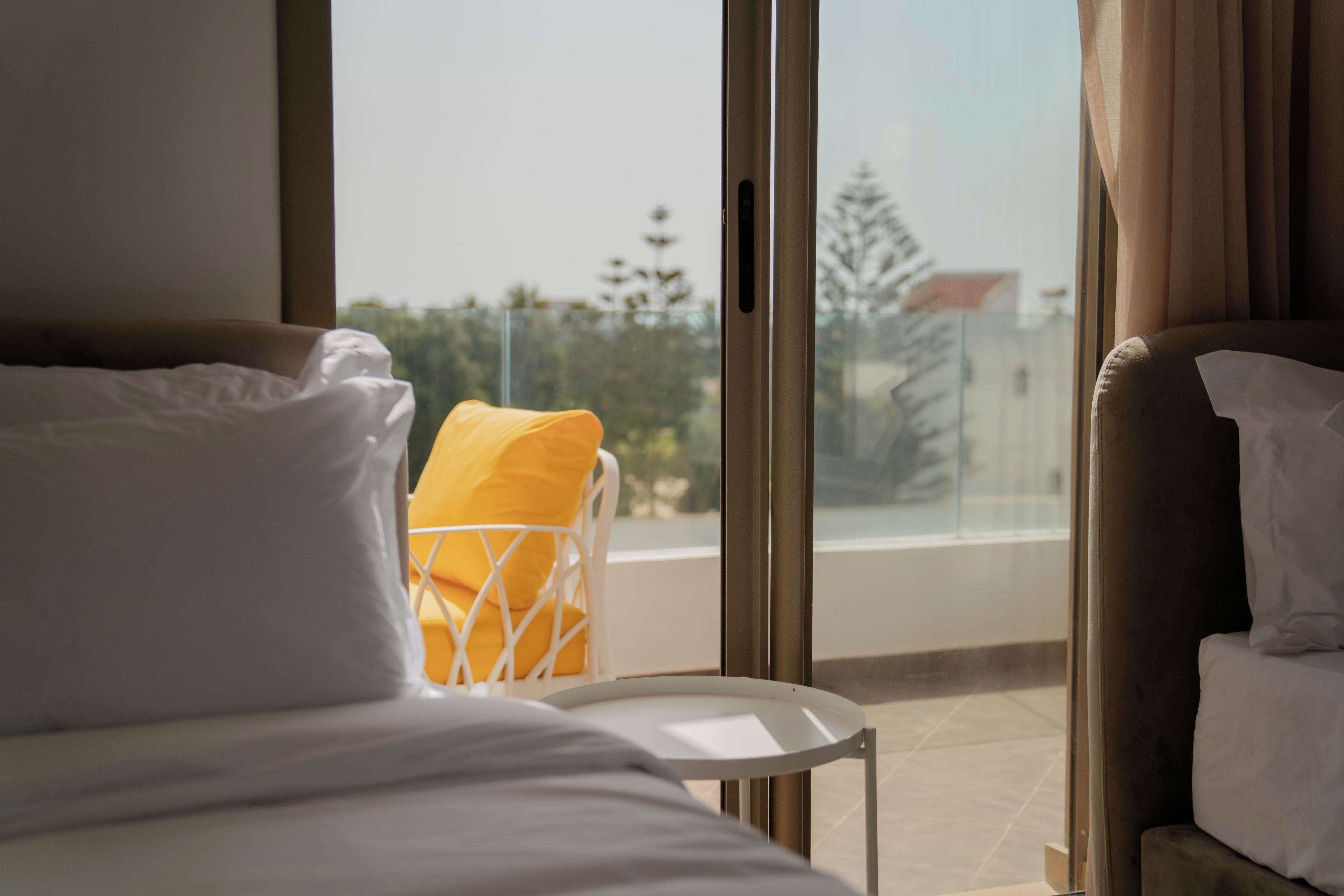 A hotel room with a bed, a chair with a yellow cushion, and a view of the outside through large windows.