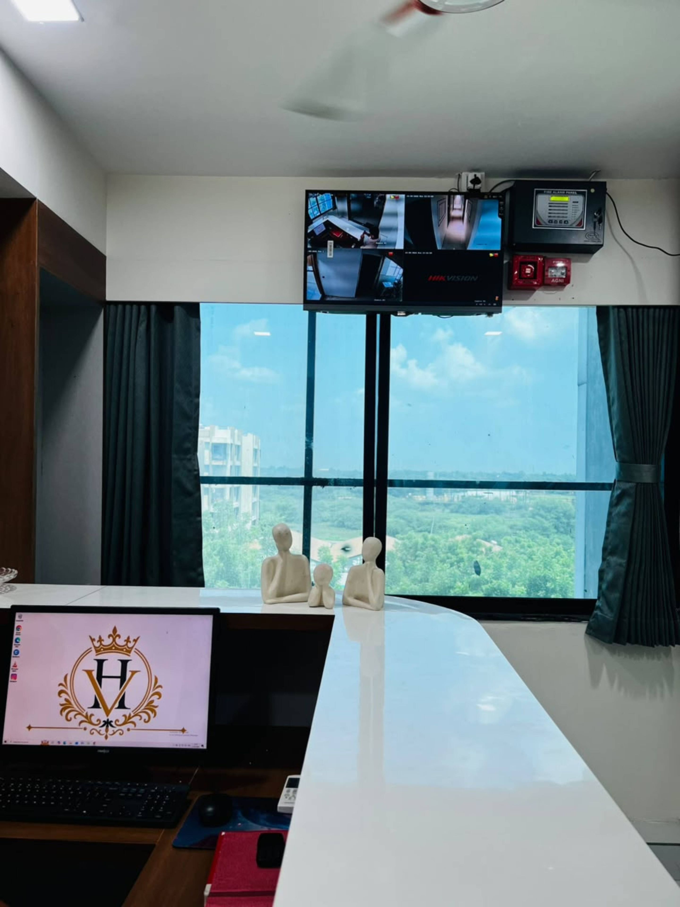 A room with a large window, a desk with a laptop, a television mounted above the desk, and a small statue on the windowsill.