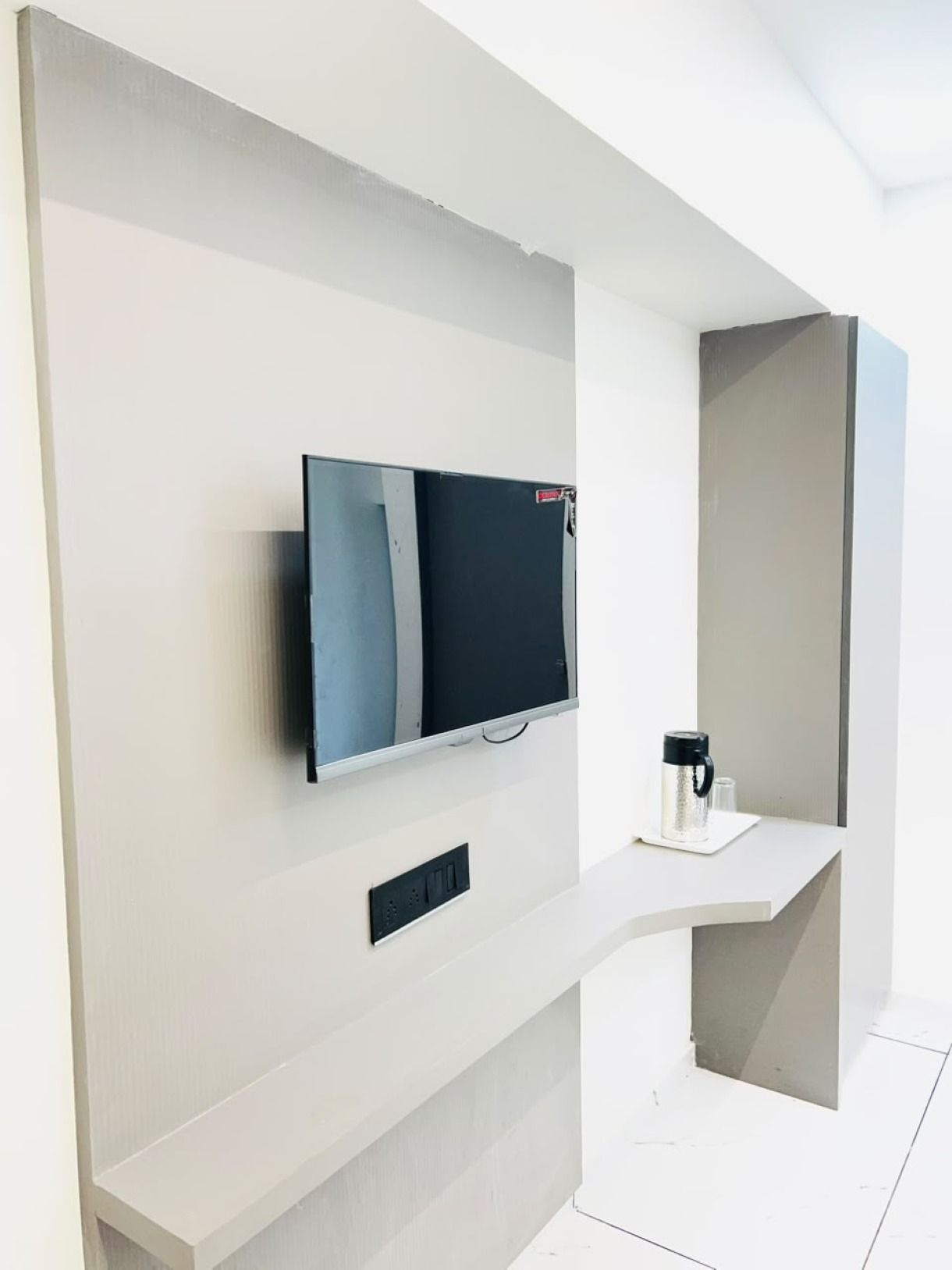 Een moderne, minimalistisch ingerichte kamer met een flatscreen televisie aan een witte muur, een klein wit tafeltje met een koffiezetapparaat en een stopcontact.