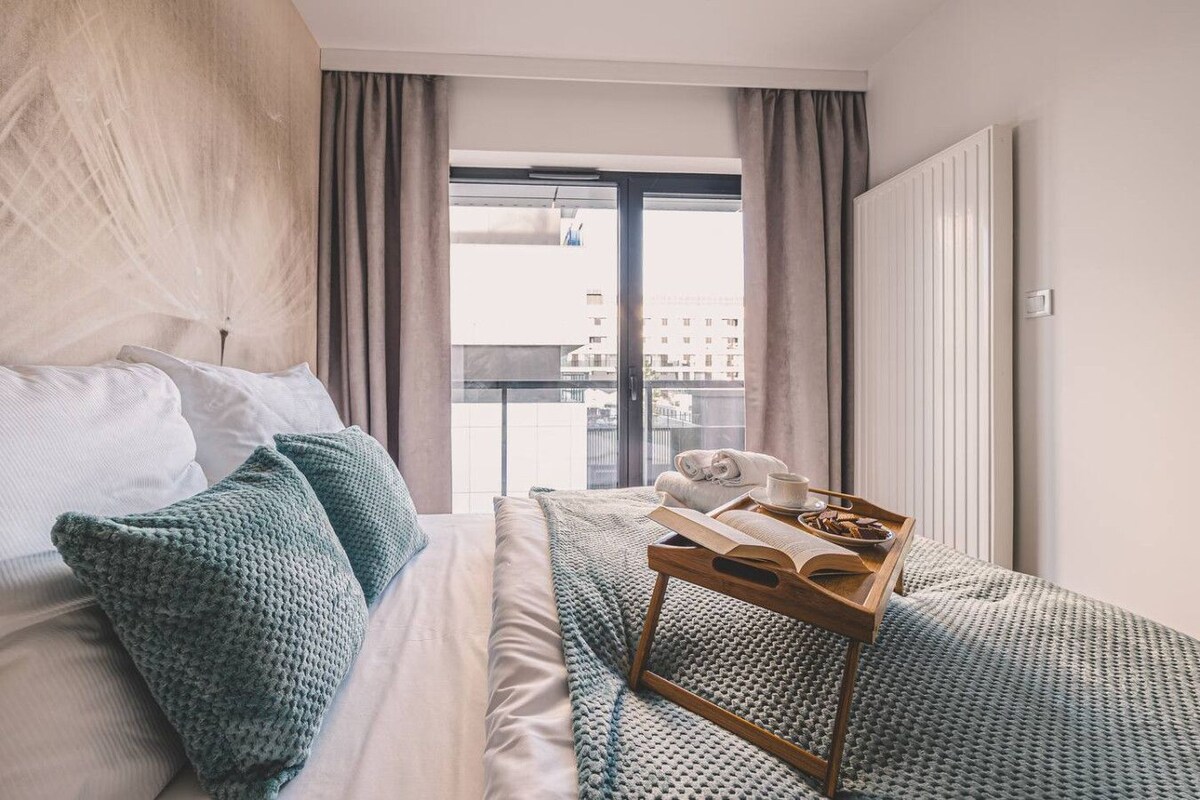 Ein Hotelzimmer mit einem Bett, einem Nachttisch mit einem Frühstücksbrettchen, einem Fenster mit Vorhängen und Blick auf Gebäude.