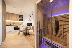 A modern kitchen with wooden cabinets, a dining area with a table and chairs, and a sauna with wooden slats and a glass door. - Rogowo Shelter by 2HomeRent (Rogowo)