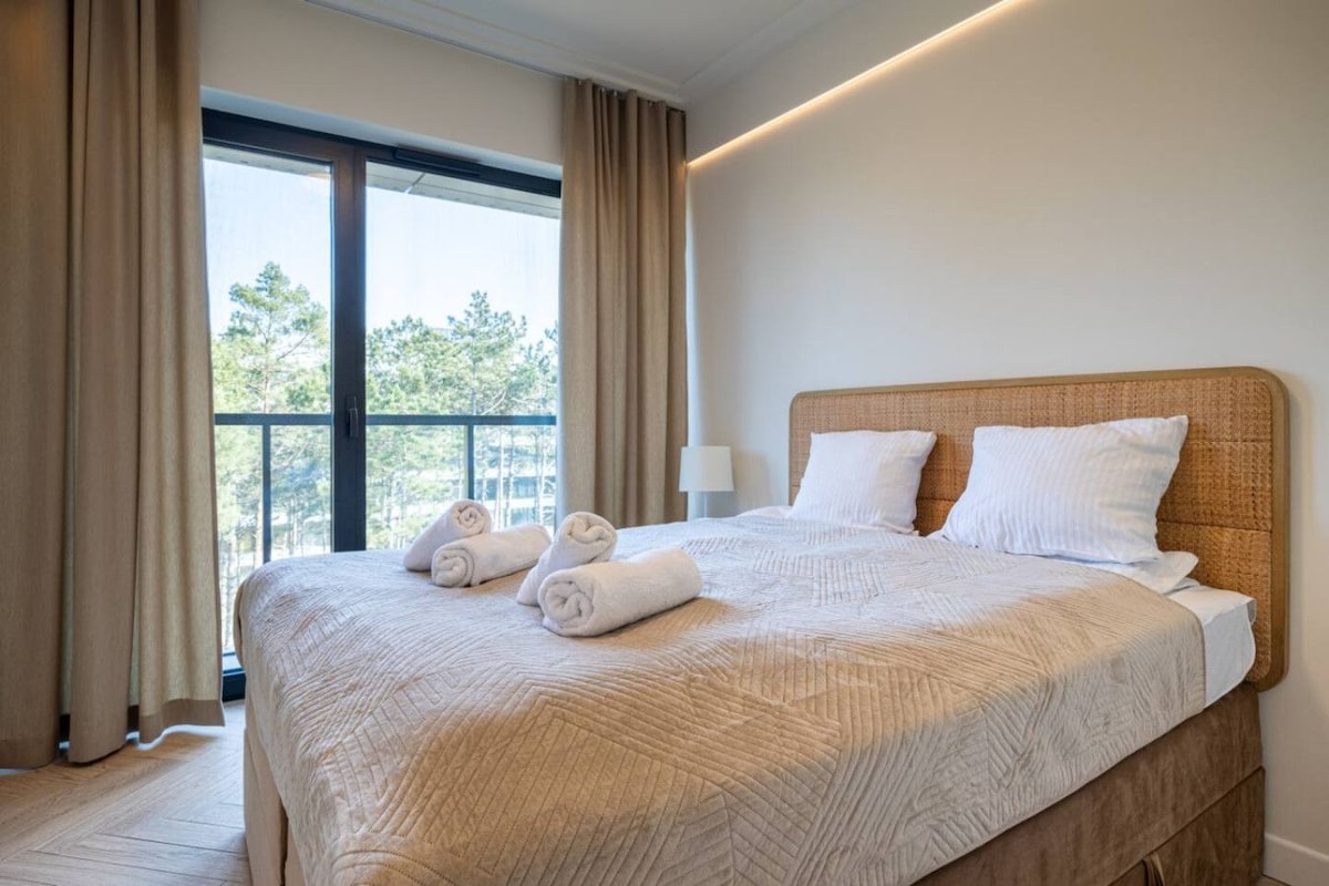 Ein Hotelzimmer mit einem großen Bett, einem Fenster mit Vorhängen und Blick auf Bäume.