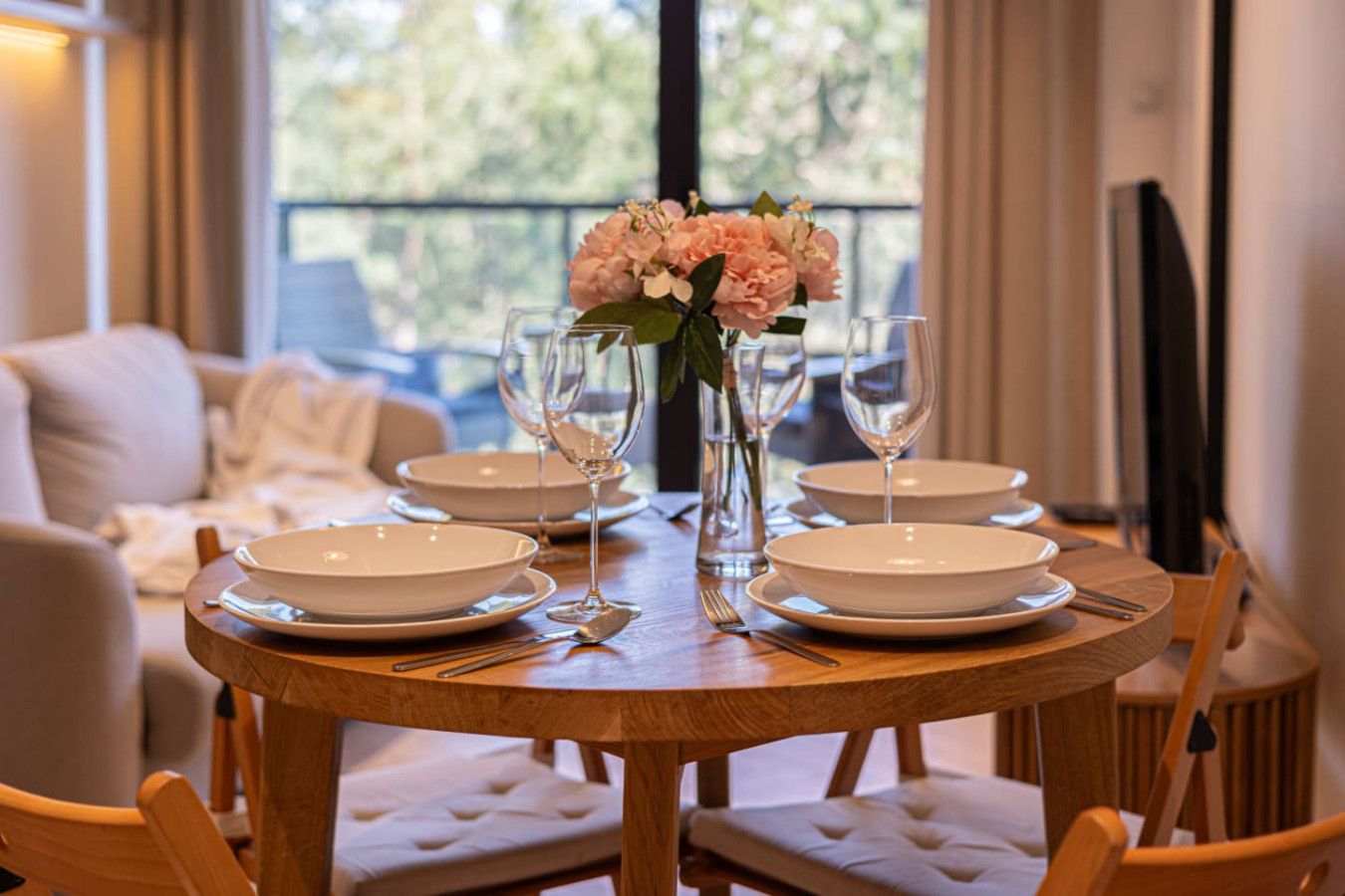 Una mesa de comedor dispuesta con platos blancos, vasos y un florero con flores, rodeada de sillas acolchadas.