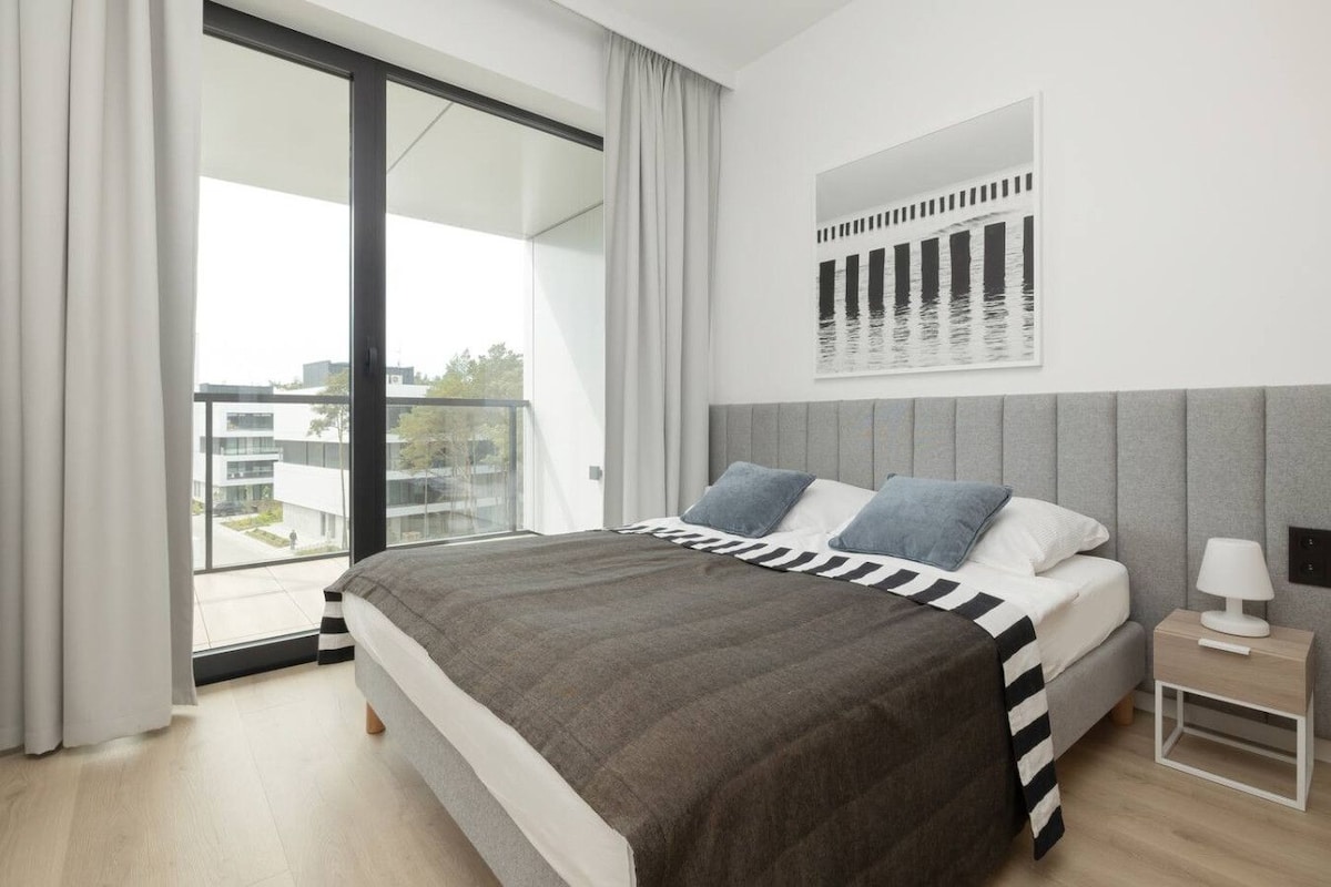 Ein modernes Schlafzimmer mit einem großen Bett, einem Nachttisch, einer Lampe und Blick auf Gebäude durch ein großes Fenster.
