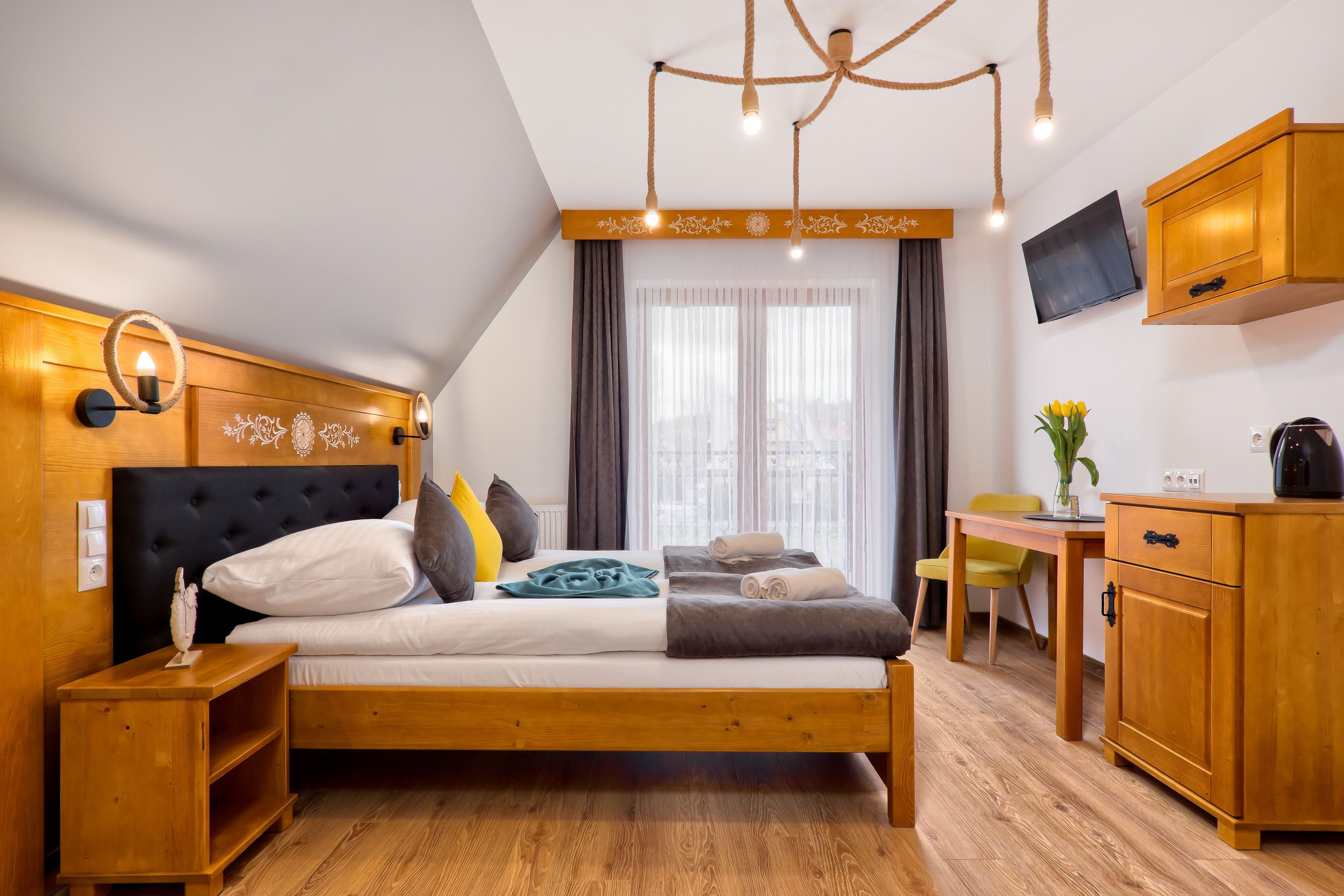 Un dormitorio moderno con cabecera de madera, cama con varios almohadones, mesita de noche, cómoda de madera, escritorio con silla y televisor en la pared.