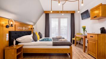 Un dormitorio moderno con cabecera de madera, cama con varios almohadones, mesita de noche, cómoda de madera, escritorio con silla y televisor en la pared.