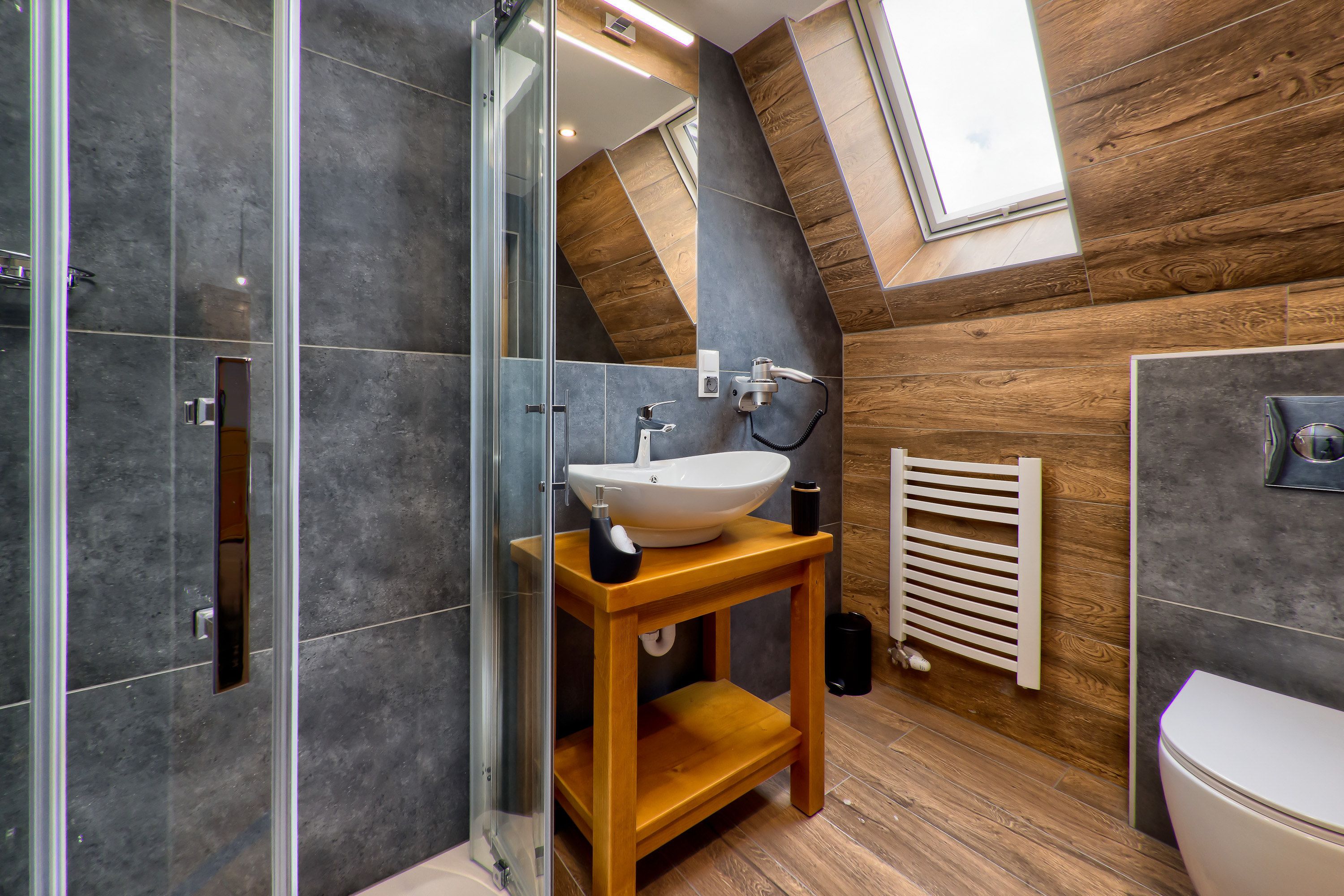 A modern bathroom with a glass shower enclosure, a wooden vanity with a sink, and a sloped ceiling with wooden paneling.