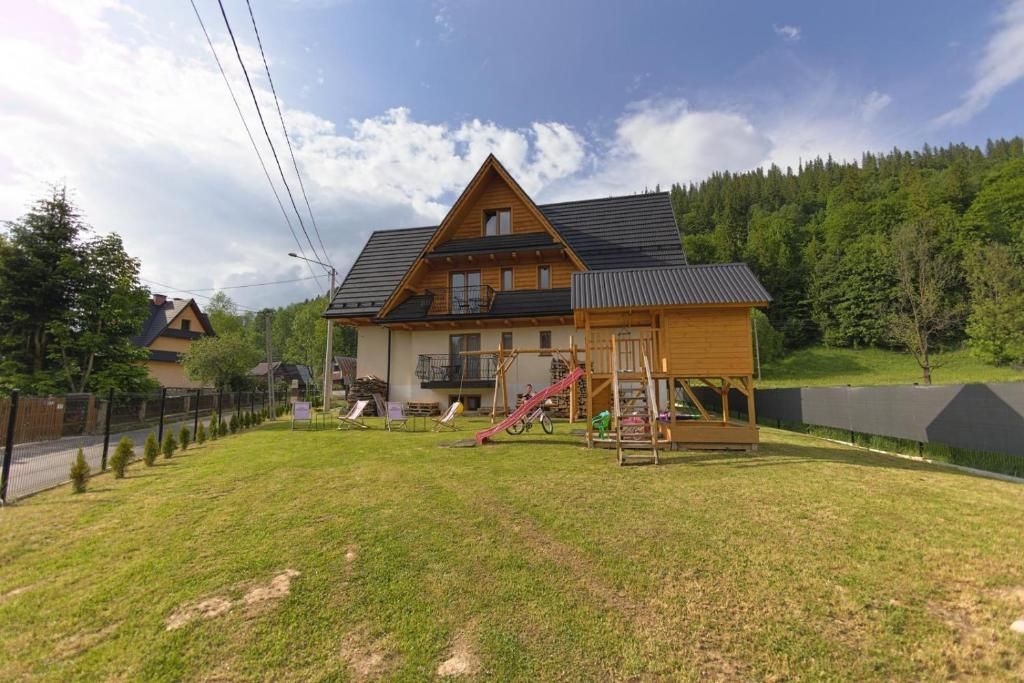 A two-story wooden house with a sloped roof, a fenced yard, and a playground with a slide.