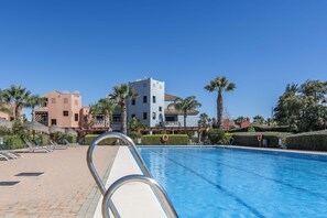 A swimming pool with lounge chairs and umbrellas, surrounded by palm trees and buildings. - El Rincon del Golf II - Isla Canela Golf GRHII54P57 (Ayamonte)