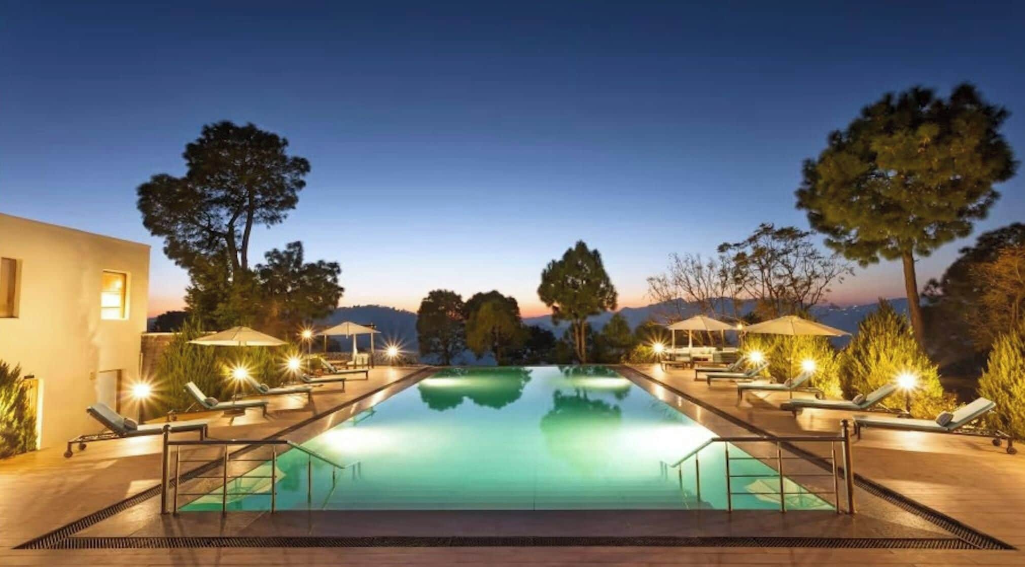 A swimming pool with lounge chairs and umbrellas, surrounded by trees and a building.