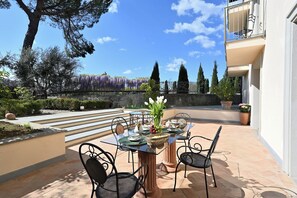 An outdoor patio with a table set for dining, surrounded by chairs, and a view of a garden with trees and purple flowers. - Your Private Tuscan Villa 17 Min to Florence · Private Pool · 6 Suites · 7 Baths (Firenze)