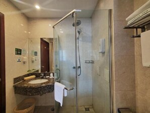Un baño moderno con ducha envuelta en vidrio, lavamanos con encimera de granito y un tozaletero a la vista.