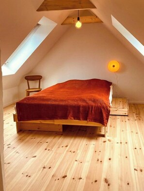 Ein Schlafzimmer im Loft-Stil mit schrägem Dach, Holzboden, einem Bett mit roter Decke, einem kleinen Holzstuhl und einer einzelnen Lampe.
