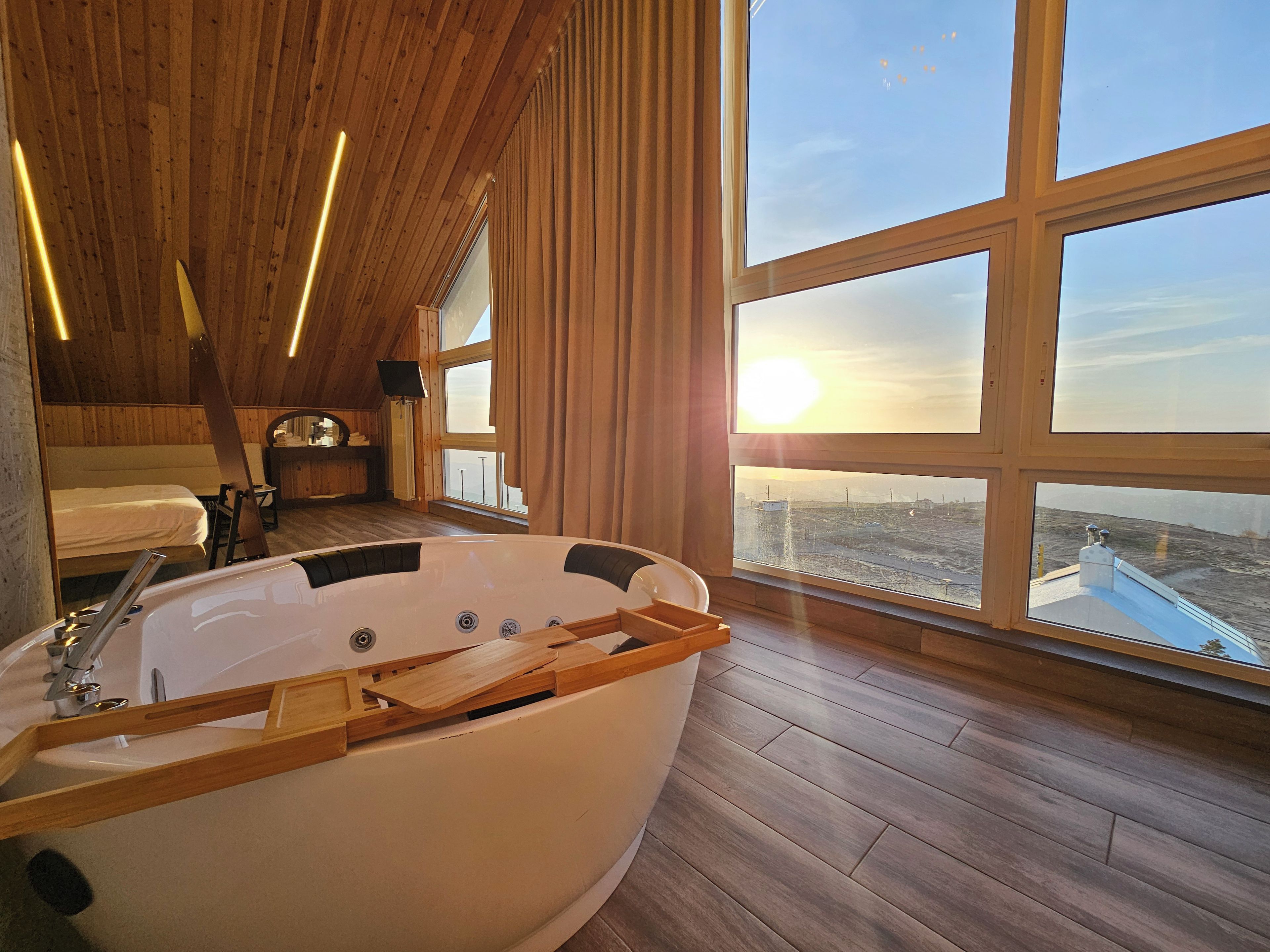 Una habitación con una bañera grande, piso de madera, una cama y vistas de la ciudad a través de amplias ventanas.