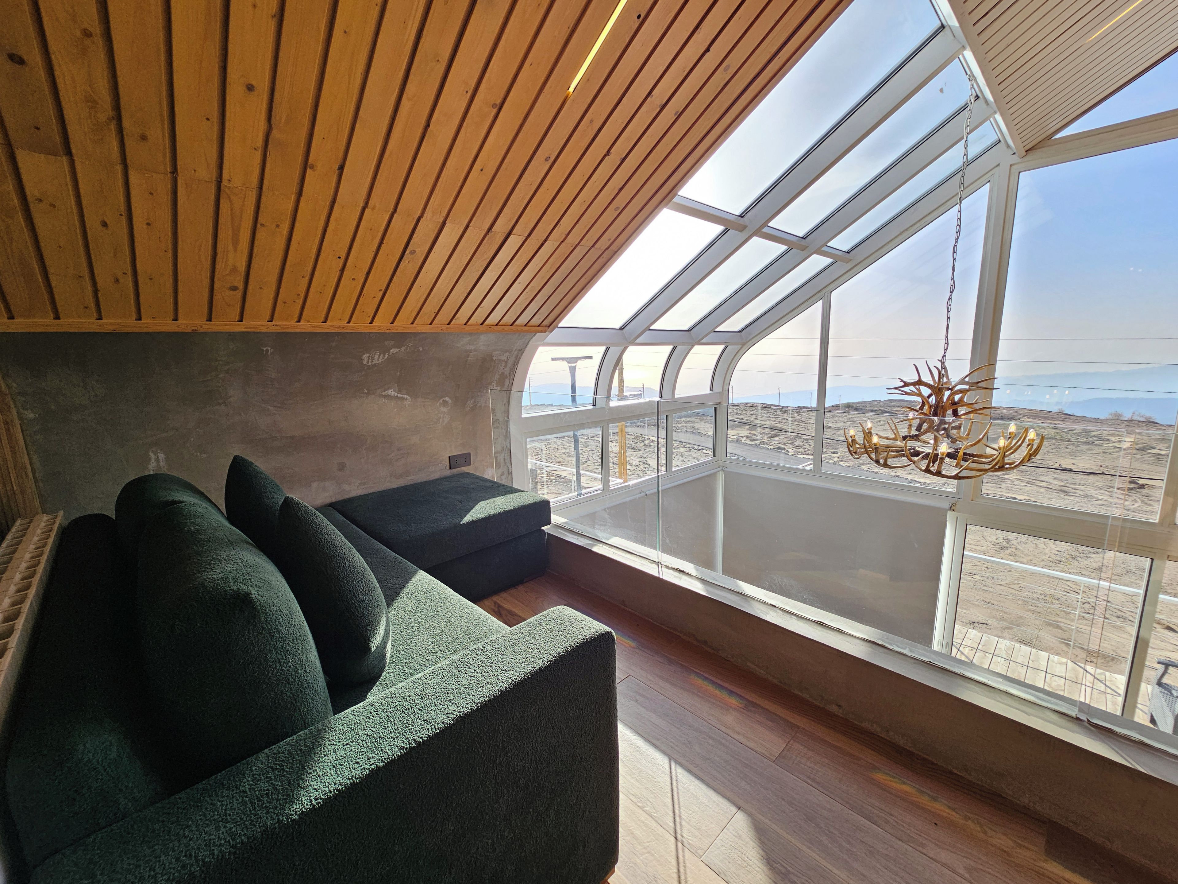 Una habitación con techos altos y techo de madera, un sofá verde con cojines, una lámpara de araña y un ventanal con vistas a un paisaje desértico.