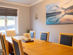 A dining area with a wooden table and chairs, a window with blinds, and a painting of a mountain landscape on the wall. - Skeabost Bungalow by Interhome (Mid Skye)