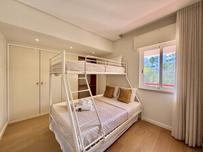 Ein Schlafraum mit Etagenbetten, einem Kleiderschrank und einem Fenster mit Vorhängen, mit Blick auf umliegende Gebäude.