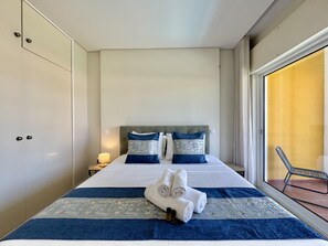 Quarto de hotel com cama, almofadas, mesa de cabeceira, janela com vista e uma cadeira do lado de fora.