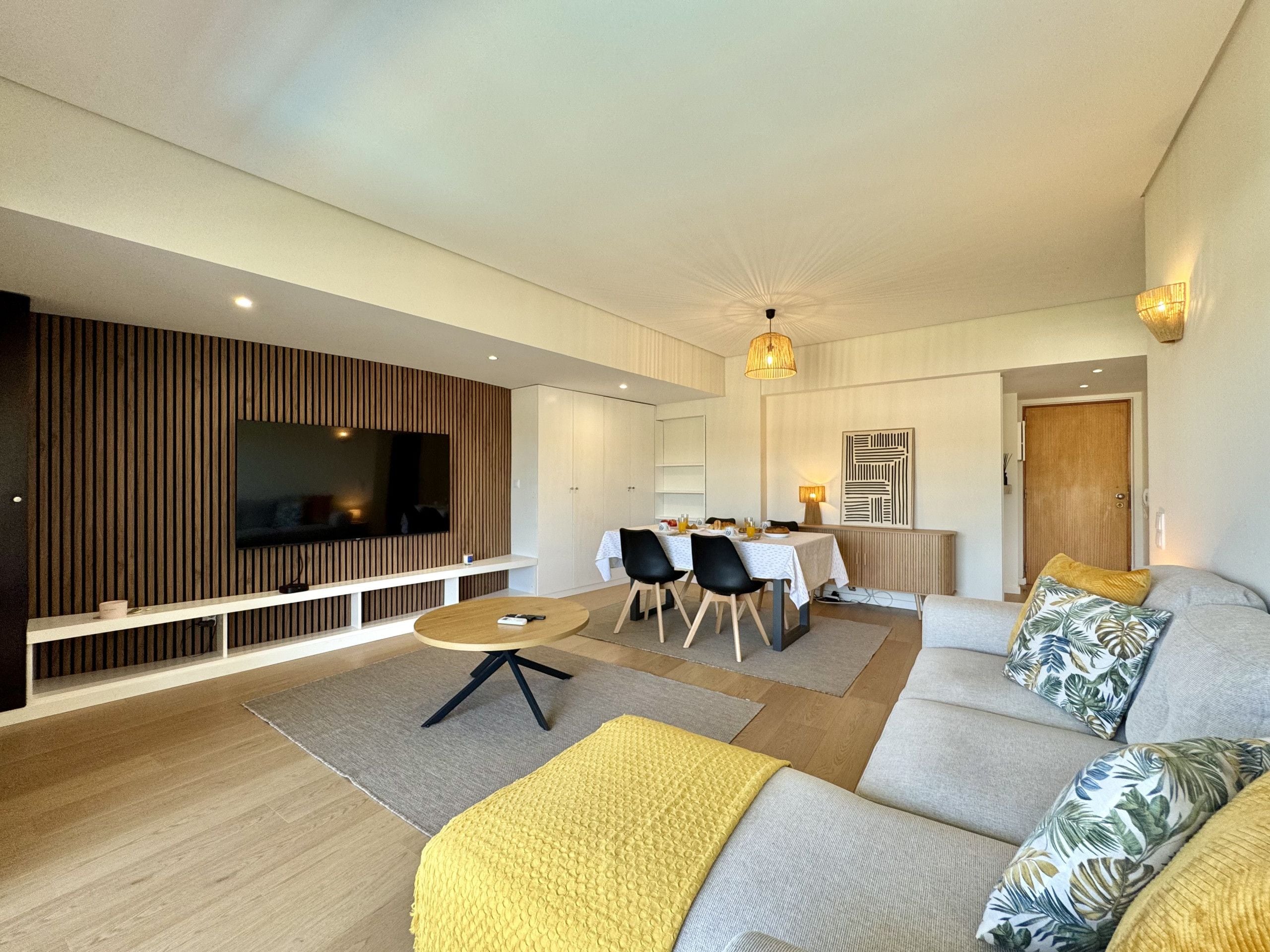 Een moderne woonkamer met een flatscreen televisie, een eethoek met tafel en stoelen, en een bank met decoratieve kussens.
