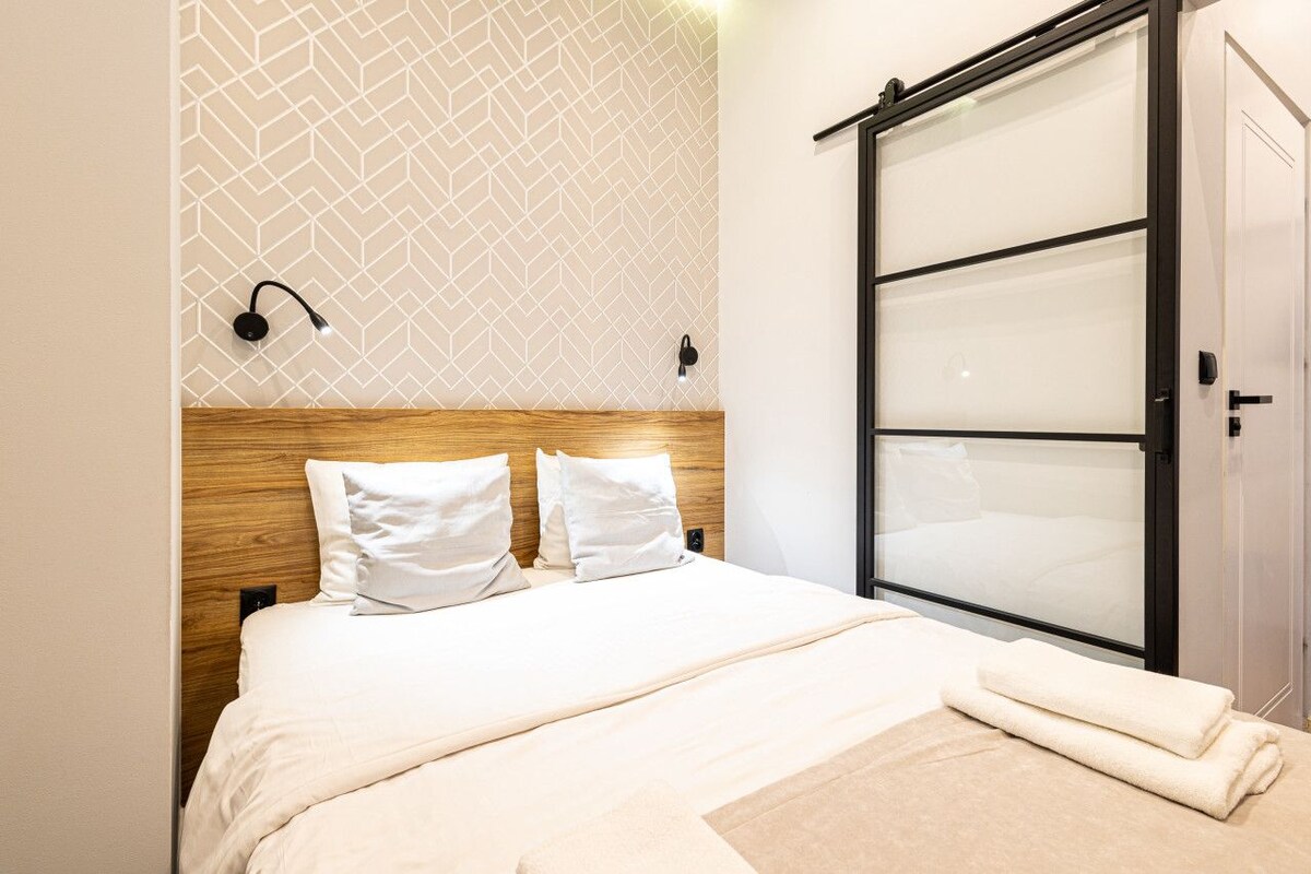 A hotel room with a bed, a wooden headboard, a geometric patterned wall, a sliding glass door, and a wall-mounted light fixture.