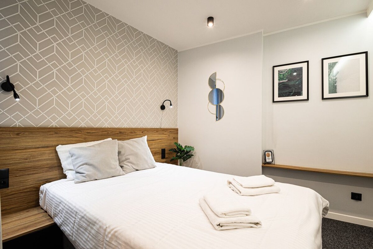 A modern hotel room with a geometric patterned wall, a wooden headboard, a bed with white linens, two framed pictures on the wall, and a bedside table with a plant.