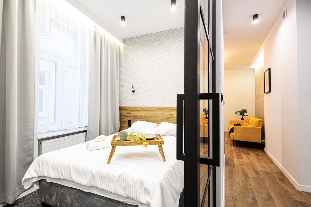 A hotel room with a bed, a wooden bedside table with a tray, a window with curtains, a wooden floor, and a yellow sofa in the adjacent living area.