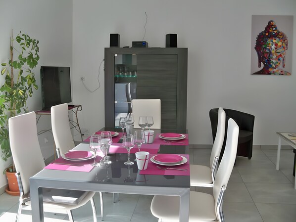 A dining area with a glass table, white chairs, pink placemats, and a modern kitchen in the background. - Cottage 'La Grange Soleil' with Private Terrace, Wi-Fi and Air Conditioning (Veigné)