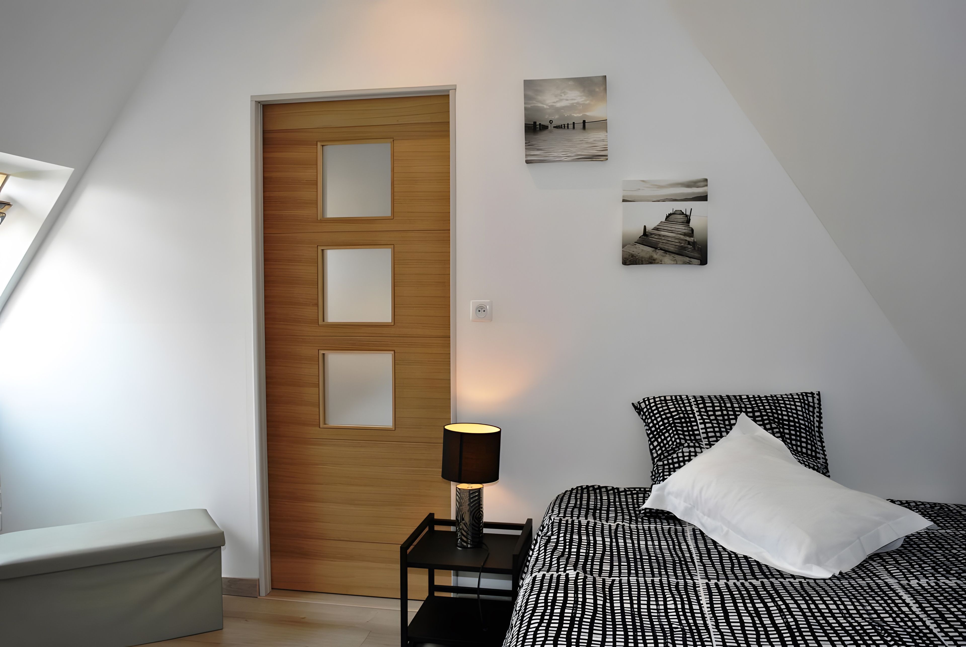 Une chambre avec une porte en bois, une table de chevet avec une lampe, un lit recouvert d’une couvre-lit à motifs noir et blanc, et deux tableaux accrochés au mur.