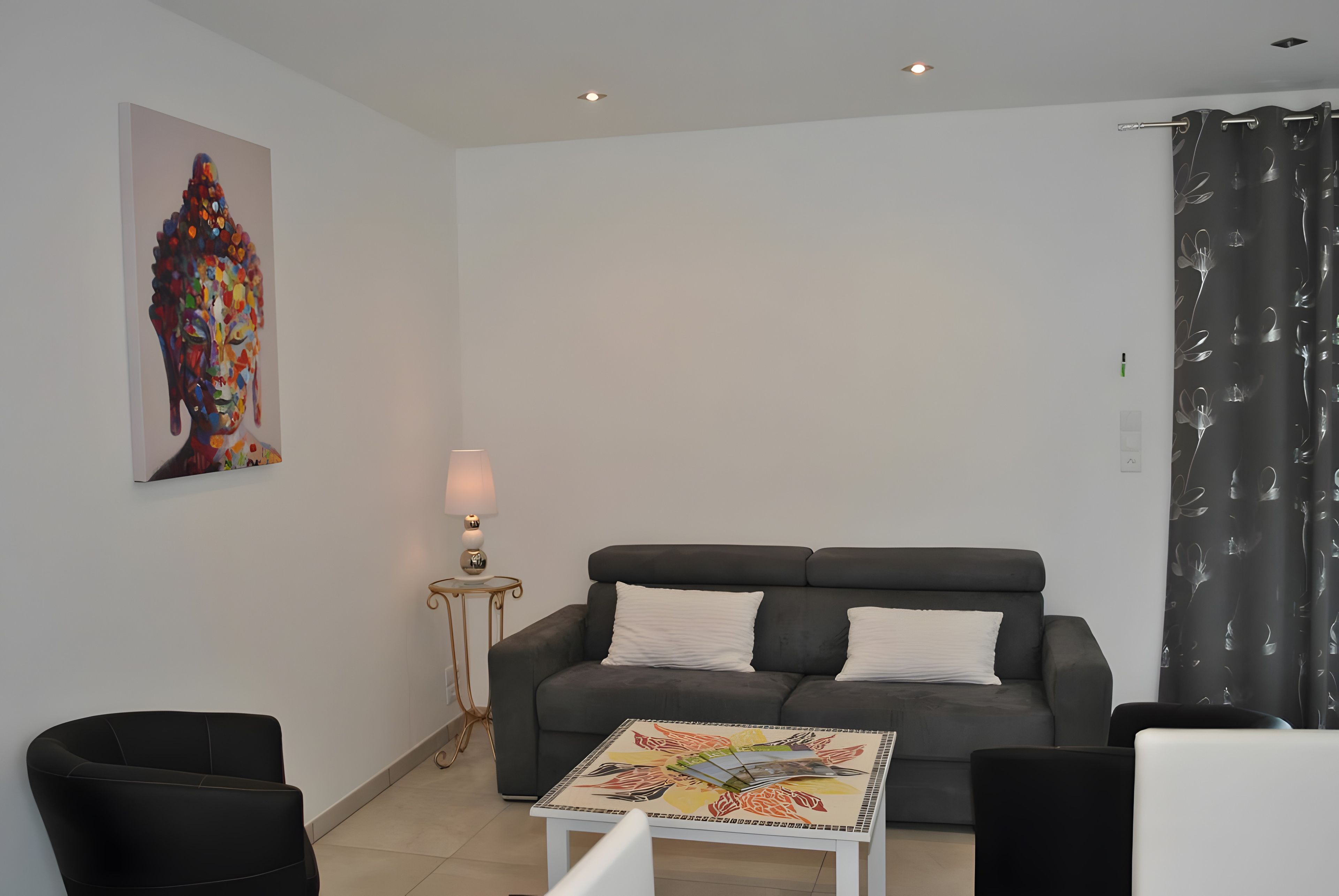 A living room with a grey sofa, two white pillows, a small table with a colorful table runner, a lamp on a side table, and a painting on the wall.