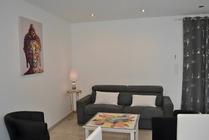 A living room with a grey sofa, two white pillows, a small table with a colorful table runner, a lamp on a side table, and a painting on the wall. - Cottage 'La Grange Soleil' with Private Terrace, Wi-Fi and Air Conditioning (Veigné)
