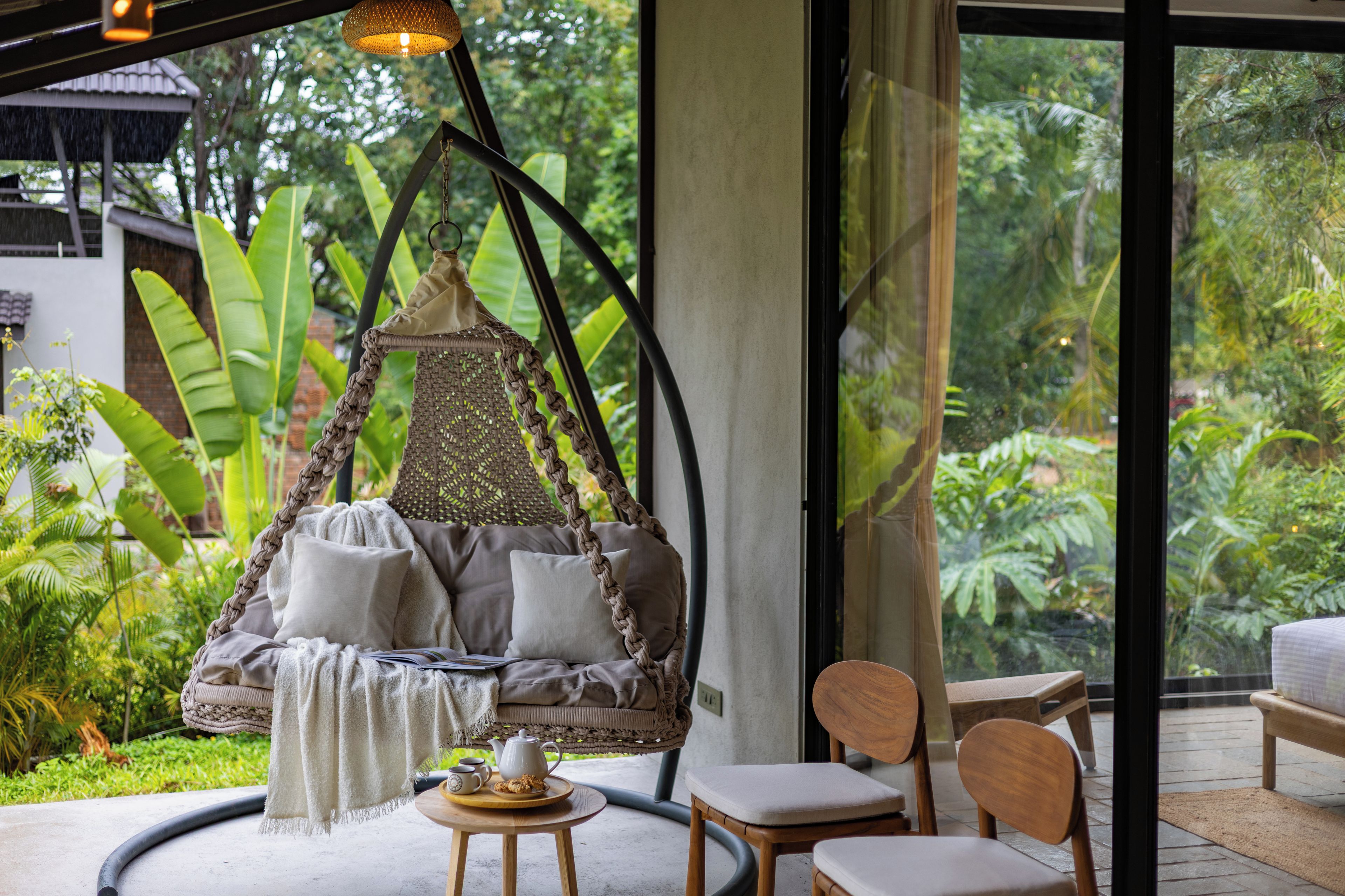 Un fauteuil suspendu avec des coussins, une table avec une théière et des biscuits, et une vue sur une végétation luxuriante à travers de grandes fenêtres.