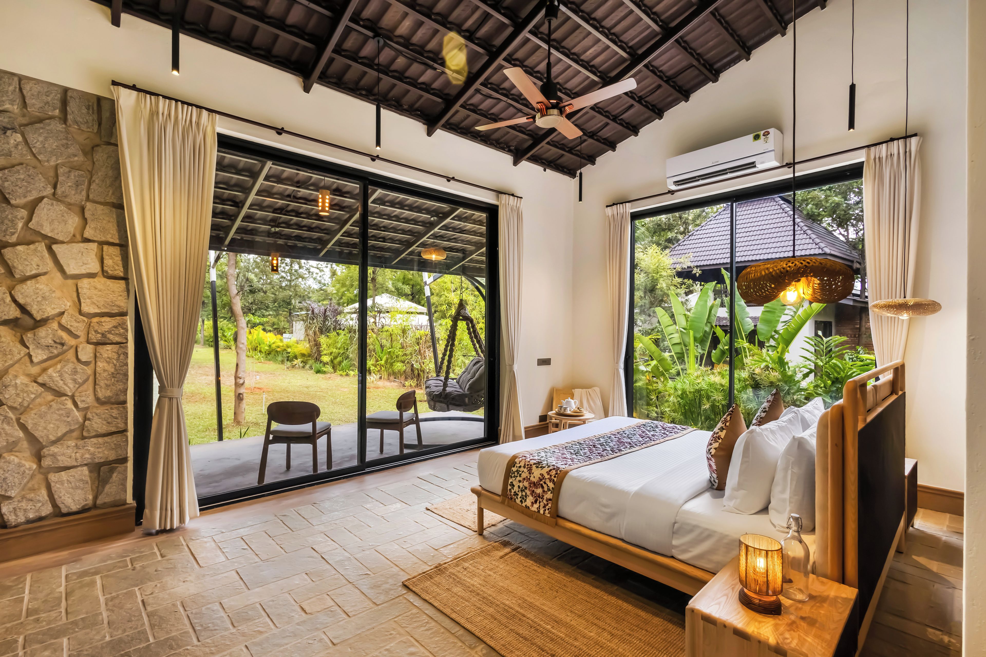 A bedroom with a bed, a nightstand, a lamp, and a view of the garden through large windows.